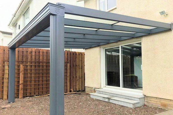 A gray metal patio cover or pergola attached to the side of a house over a set of steps leading to a sliding glass door.