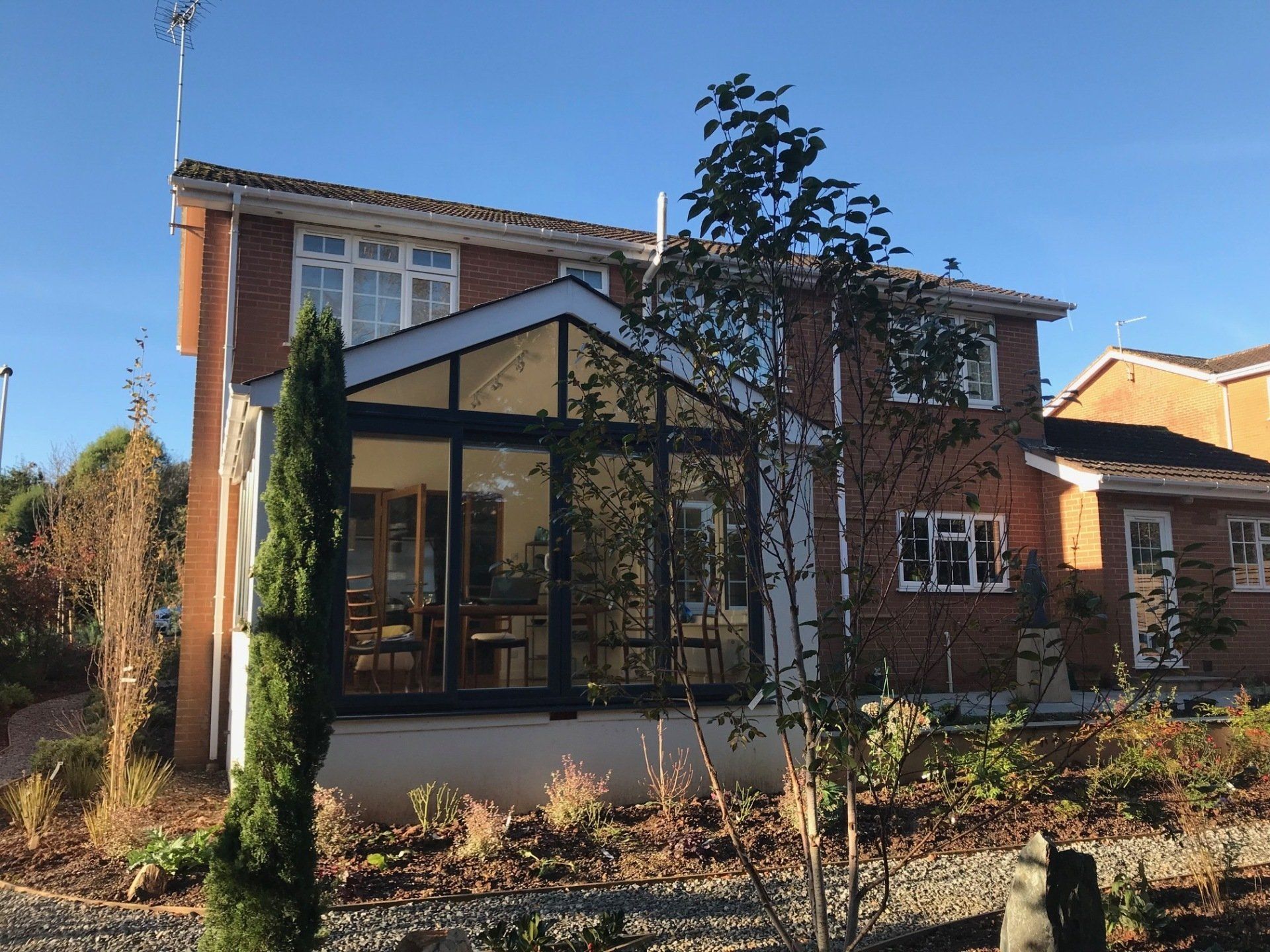 A two-story brick house with a dark-framed glass sunroom extension under a clear blue sky, surrounded by garden beds.