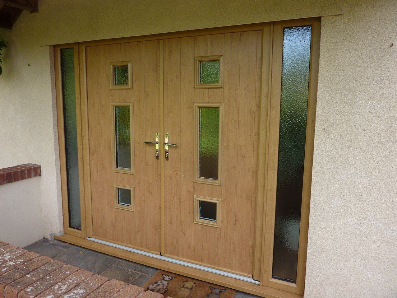 Light oak double entry doors with vertical glass panels and two matching side lights set in a textured cream wall.
