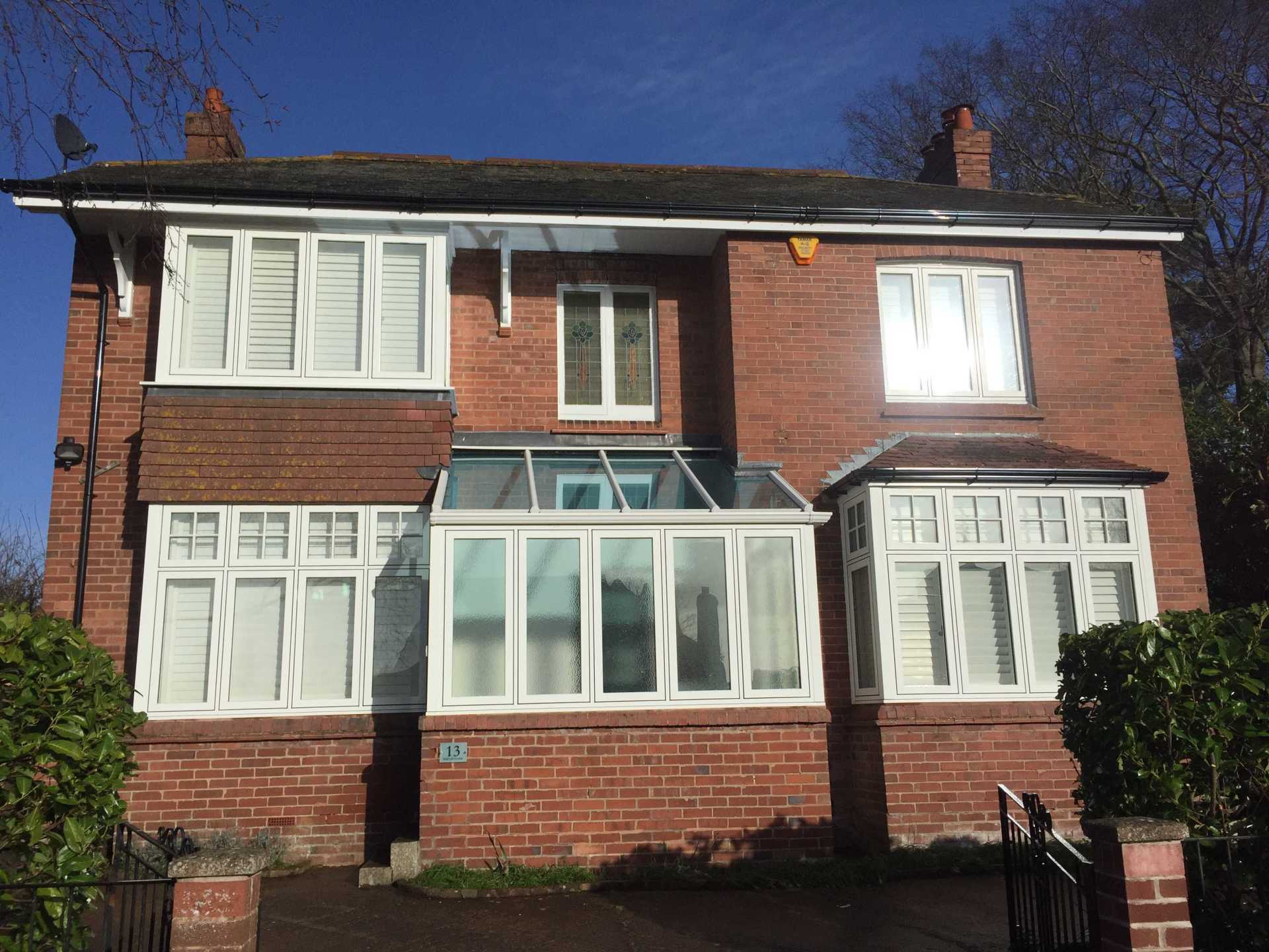 A two-story red brick house with white window frames, a glass conservatory, and green bushes against a clear blue sky.