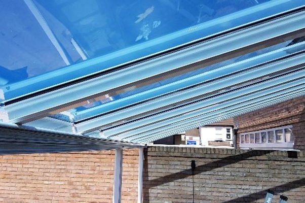 A low-angle view of a glass conservatory roof with white framing, attached to a brick wall under a bright blue sky.