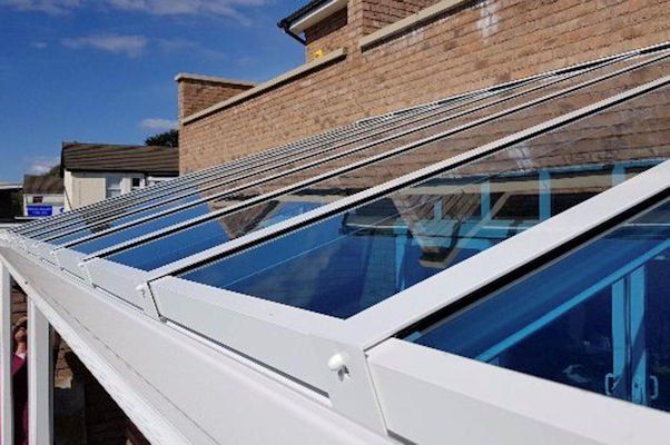 A white-framed conservatory roof with blue-tinted glass panels attached to a brick house under a blue sky.