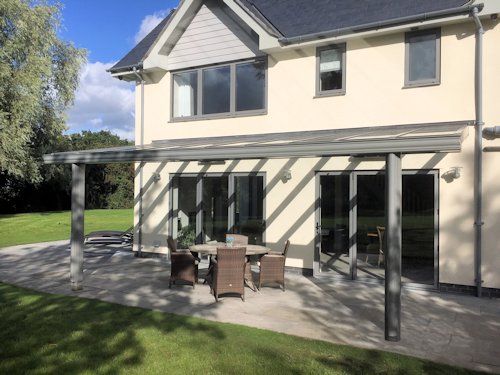 A light-colored house with a patio featuring a modern metal pergola over a table and chair set on a sunny day.