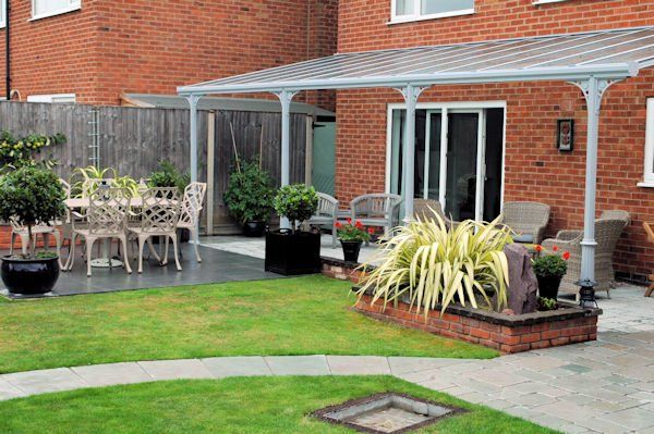 A brick house patio features a covered seating area with wicker furniture, a small lawn, and a stone garden bed.