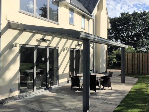 A light-colored house features a modern grey patio cover over a stone terrace with outdoor dining furniture.