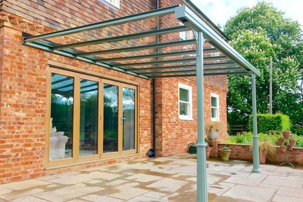 A glass-roofed metal patio canopy attached to a brick house with large folding glass doors and a stone-paved patio.