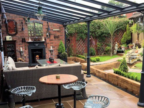 A cozy outdoor covered patio with a brick fireplace, wicker seating, and metal tractor-seat stools overlooking a garden.
