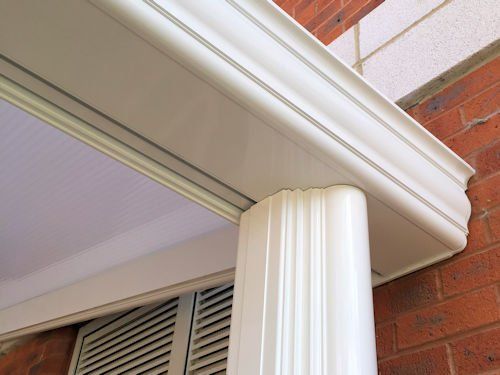 A close-up view of a white, fluted porch column supporting a decorative white gutter system against a red brick wall.