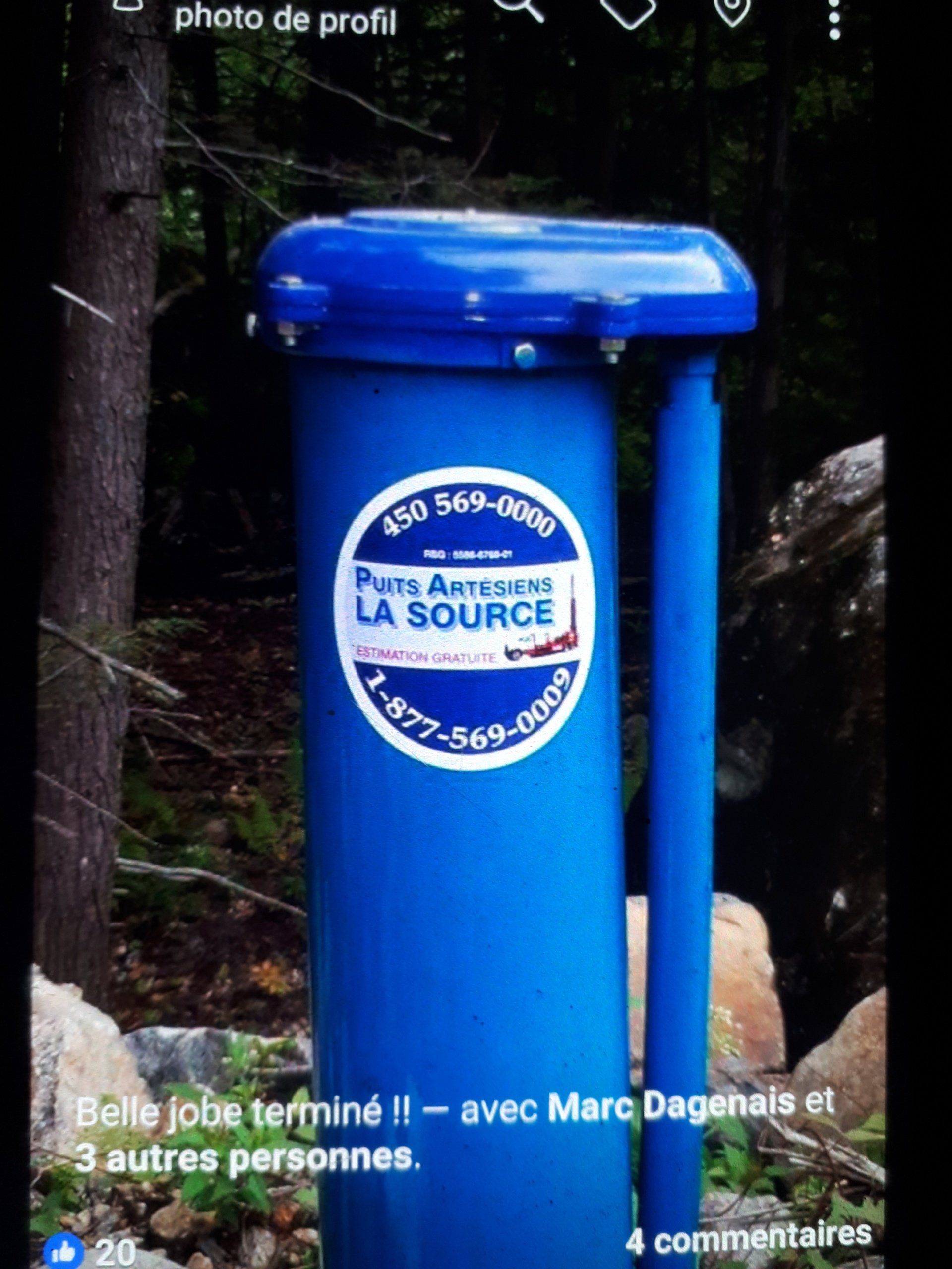 Blue recycling bin with a white circular logo, outdoors in a wooded area