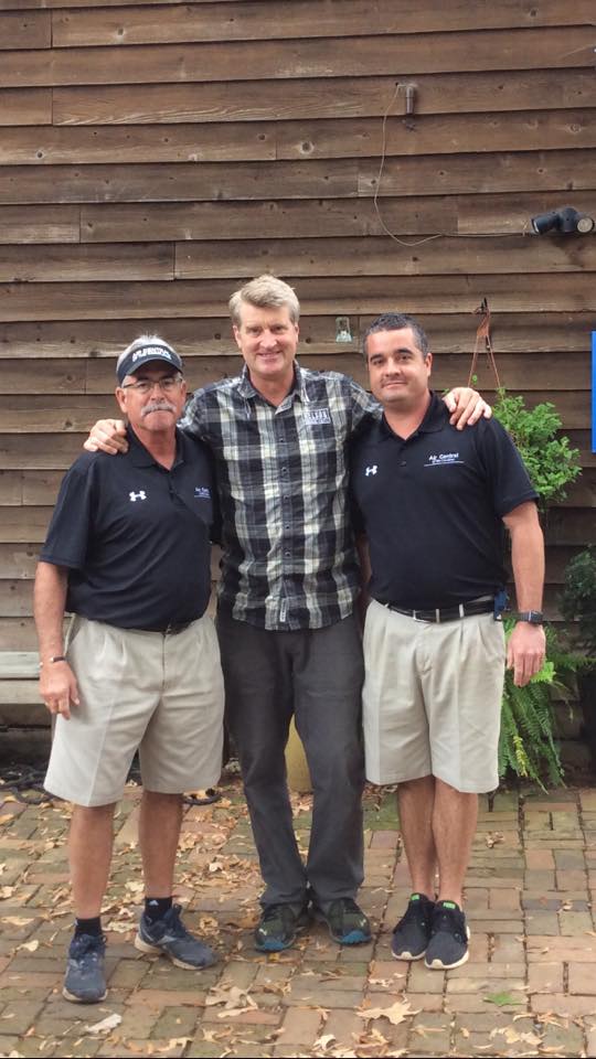 Three men standing outdoors; middle man has arms around the other two. Wooden wall background.