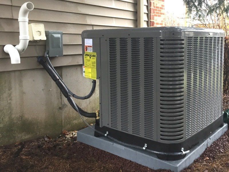 Central air conditioning unit on a concrete base next to a building. Wiring and pipes are visible.