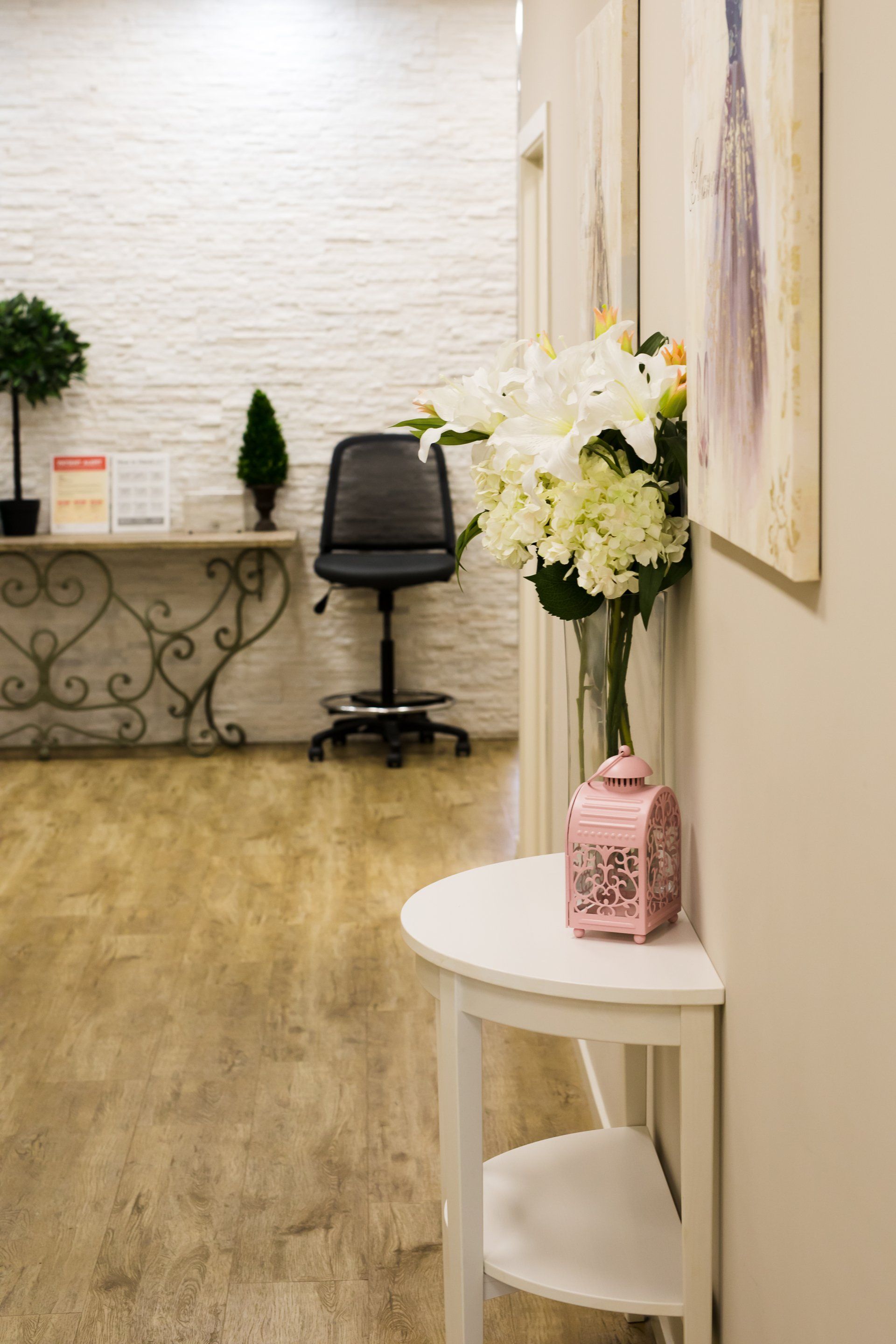 Small office waiting area with desk, black chair, floral arrangement, and white side table in a beige hallway.