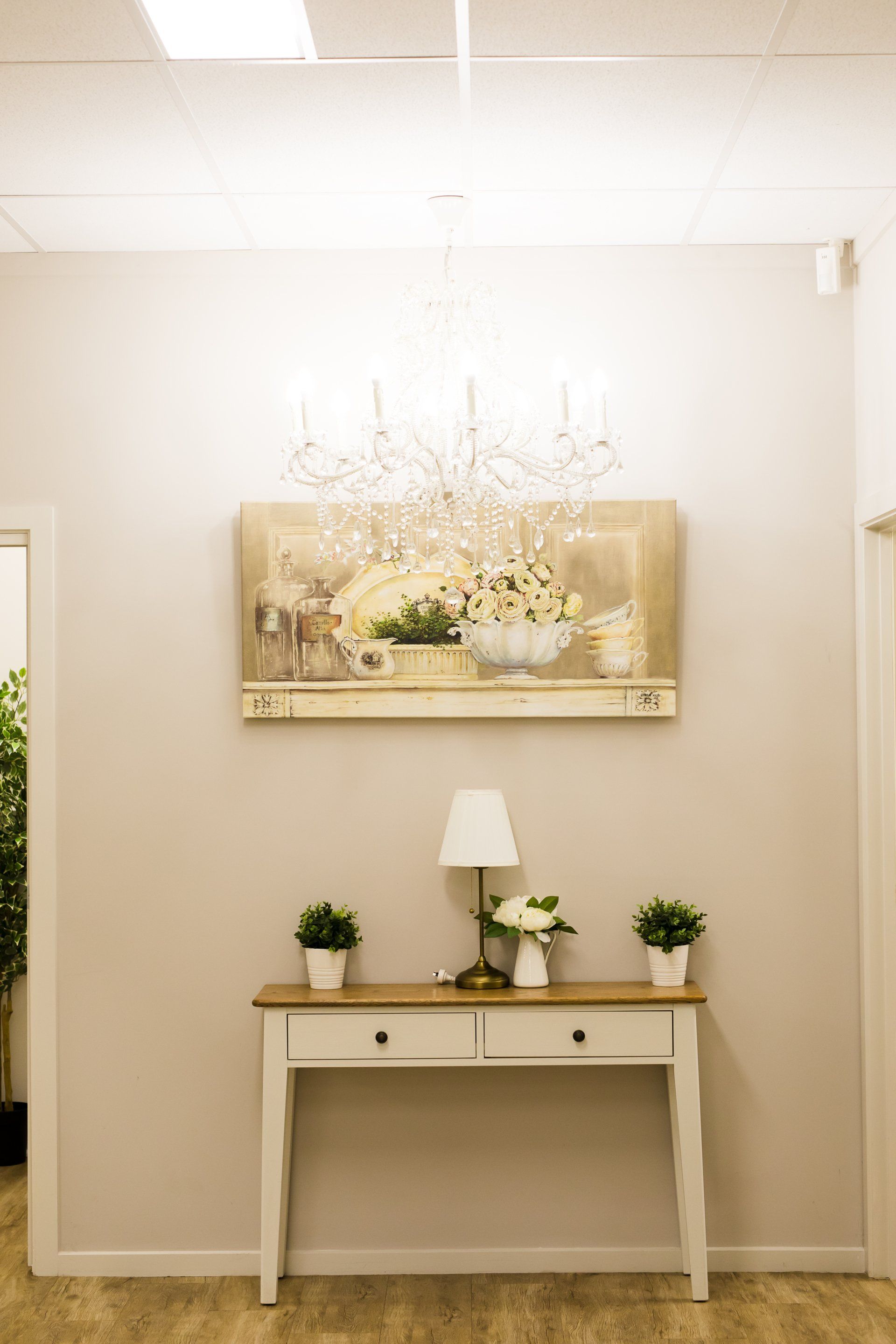 Elegant hallway with a white console table, flowers, lamp, and chandelier beneath framed wall art.