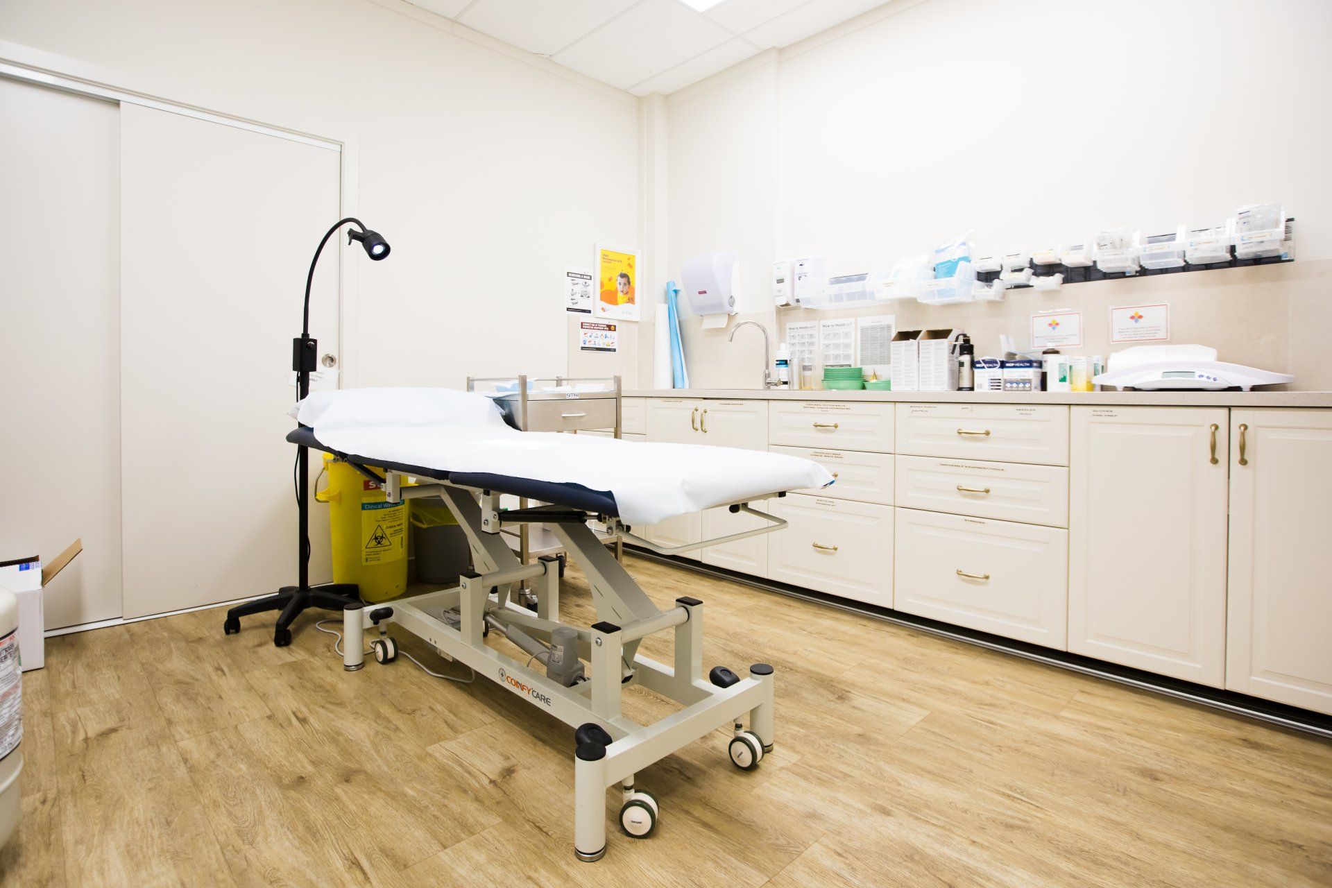 Empty medical exam room with exam table, cabinets, and equipment on wood flooring