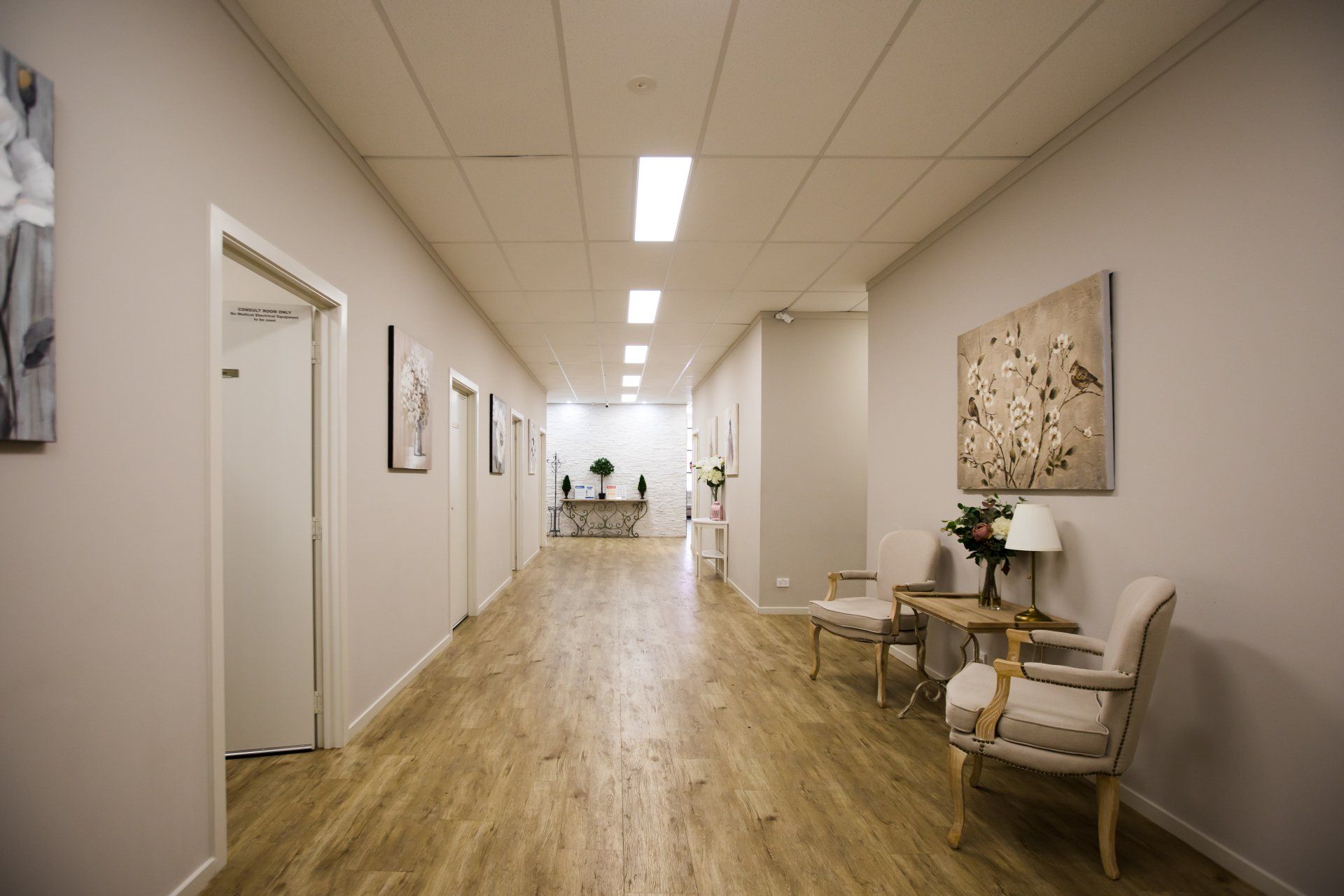 Bright office hallway with wood floors, white walls, doors, framed art, and seating area at the end.