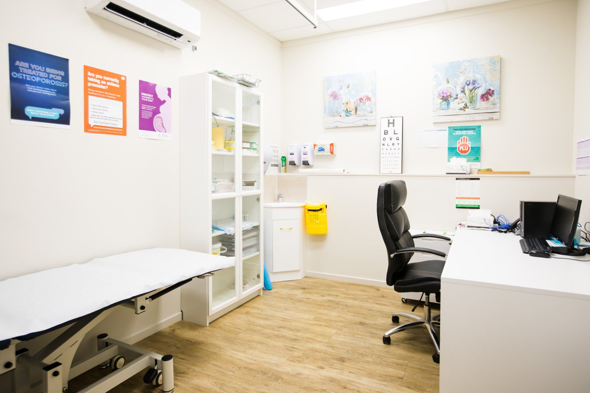 Bright medical exam room with exam table, desk, chair, wall charts, and cabinets