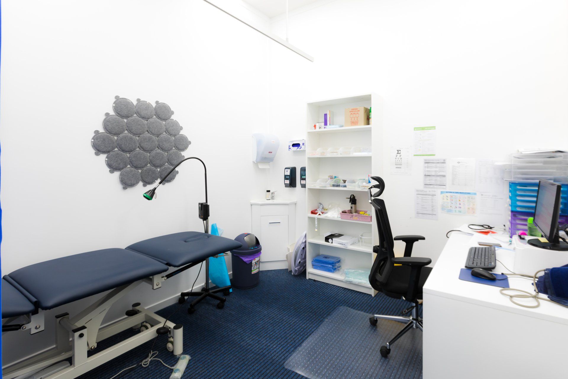 Modern medical exam room with treatment table, desk, shelves, and office chair