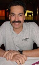 A man with a mustache is sitting at a table with his hands folded.
