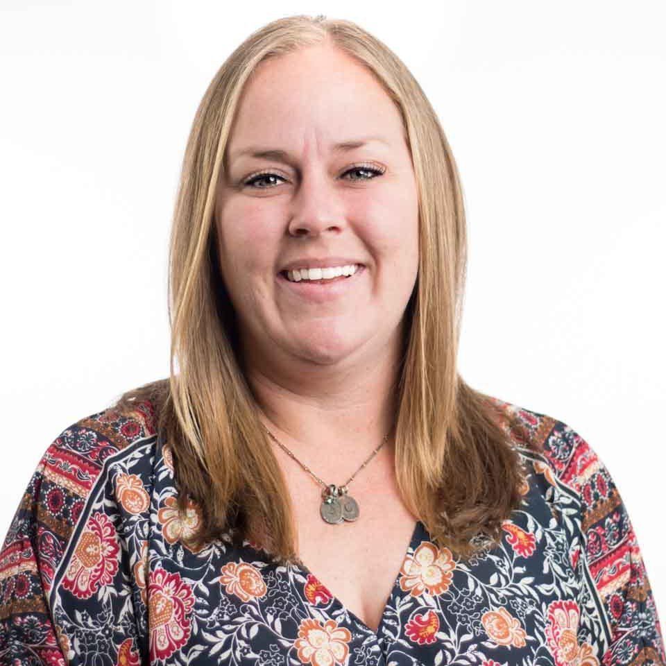 Smiling person with straight blonde hair wearing a patterned blouse and necklace against a white background