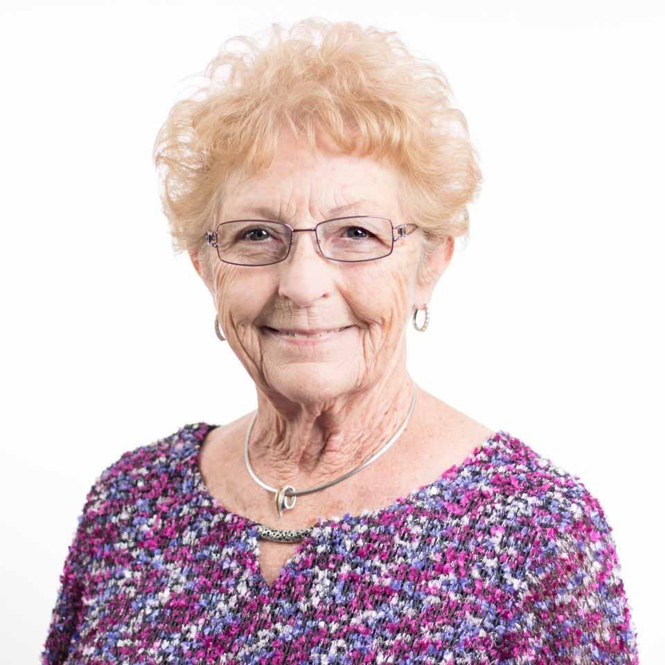 Smiling woman with short curly blonde hair and glasses, wearing a purple patterned top.