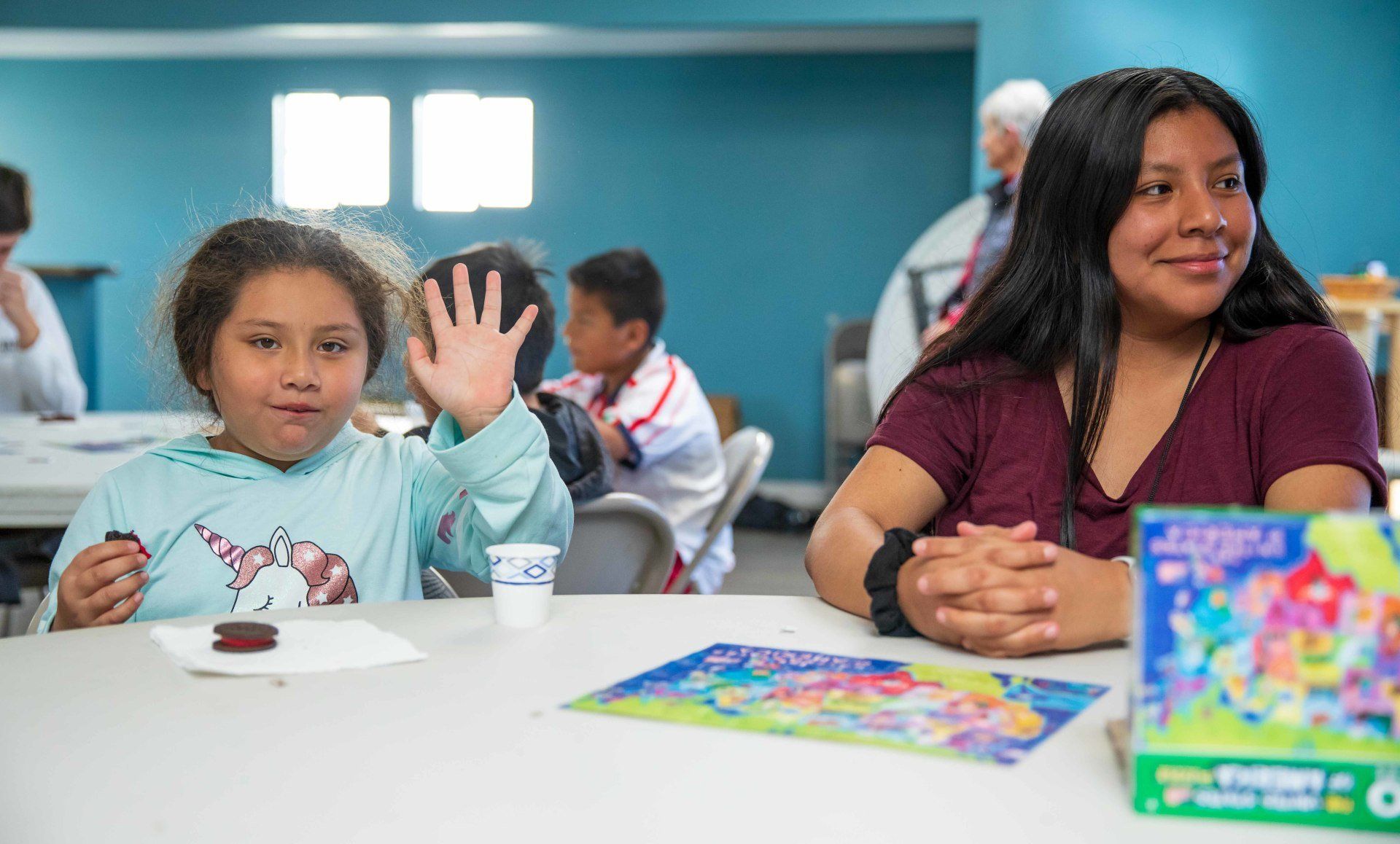 Two children at a table; one waves, with colorful books and papers in a classroom setting.