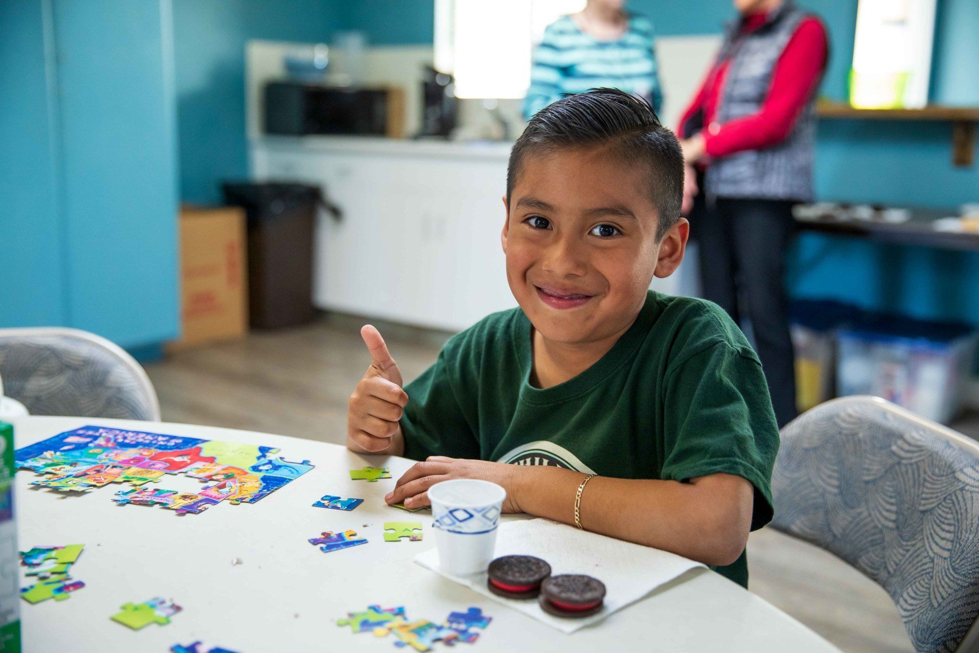 Child at a table with puzzle pieces, smiling and giving a thumbs-up in a classroom.