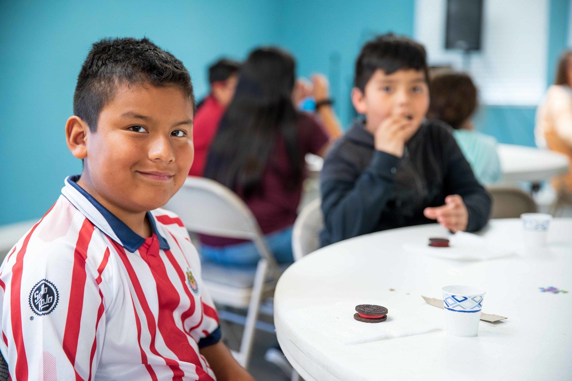 Child in a red-and-white striped shirt sitting at a table with other children in a classroom or cafeteria