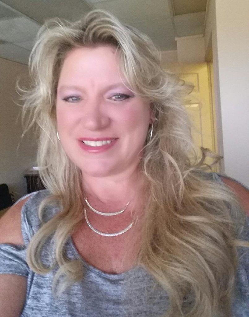 A woman wearing a gray shirt and a necklace is smiling for the camera.