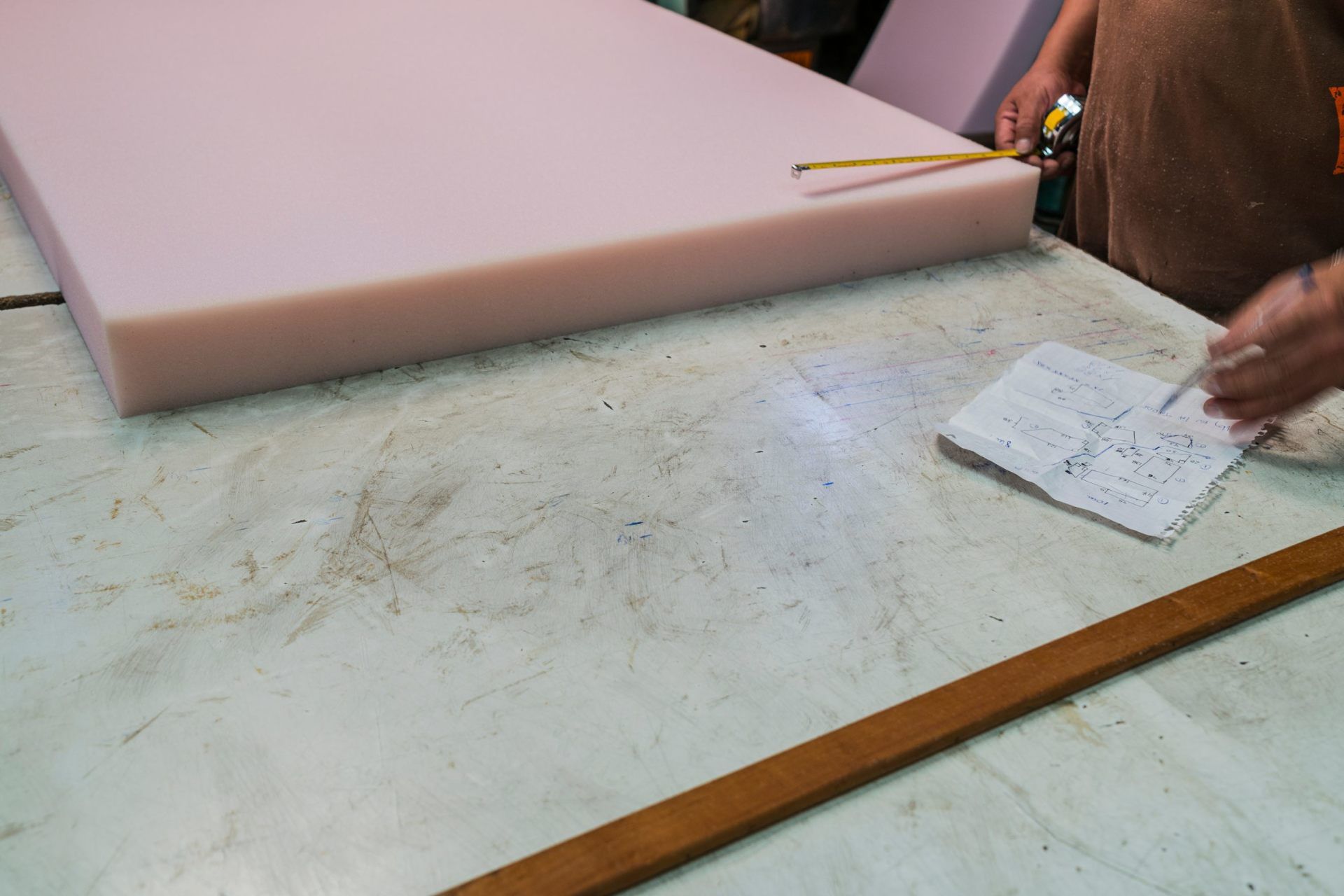 Person measuring foam with a tape measure on a workbench, looking at paper.