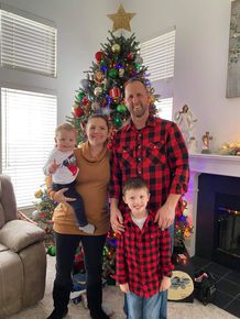 Family of four in red plaid pajamas posing in front of a decorated Christmas tree and fireplace