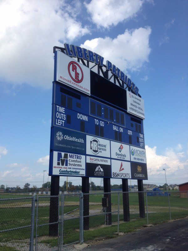 Large blue scoreboard at a baseball field under a cloudy sky, beside a chain-link fence.