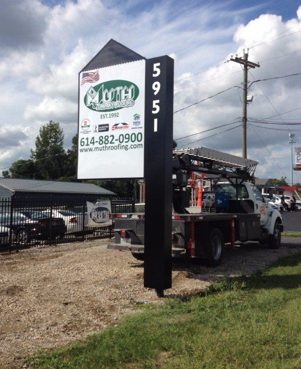 Tall business sign with green logo beside a street and parked truck under a cloudy sky.
