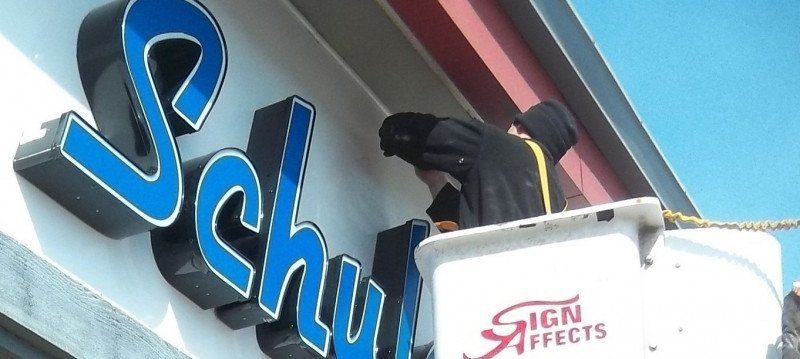 Blue “Schuh” storefront sign with a worker in a lift installing signage