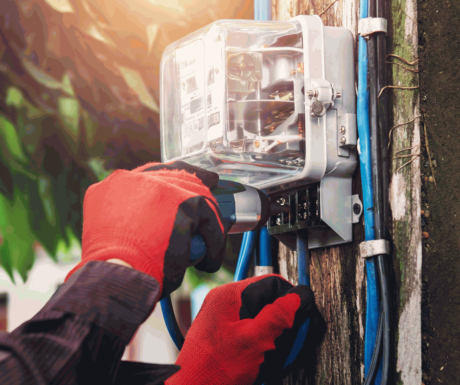 A person wearing red work gloves uses a screwdriver to service an electrical meter mounted on a wooden utility pole.