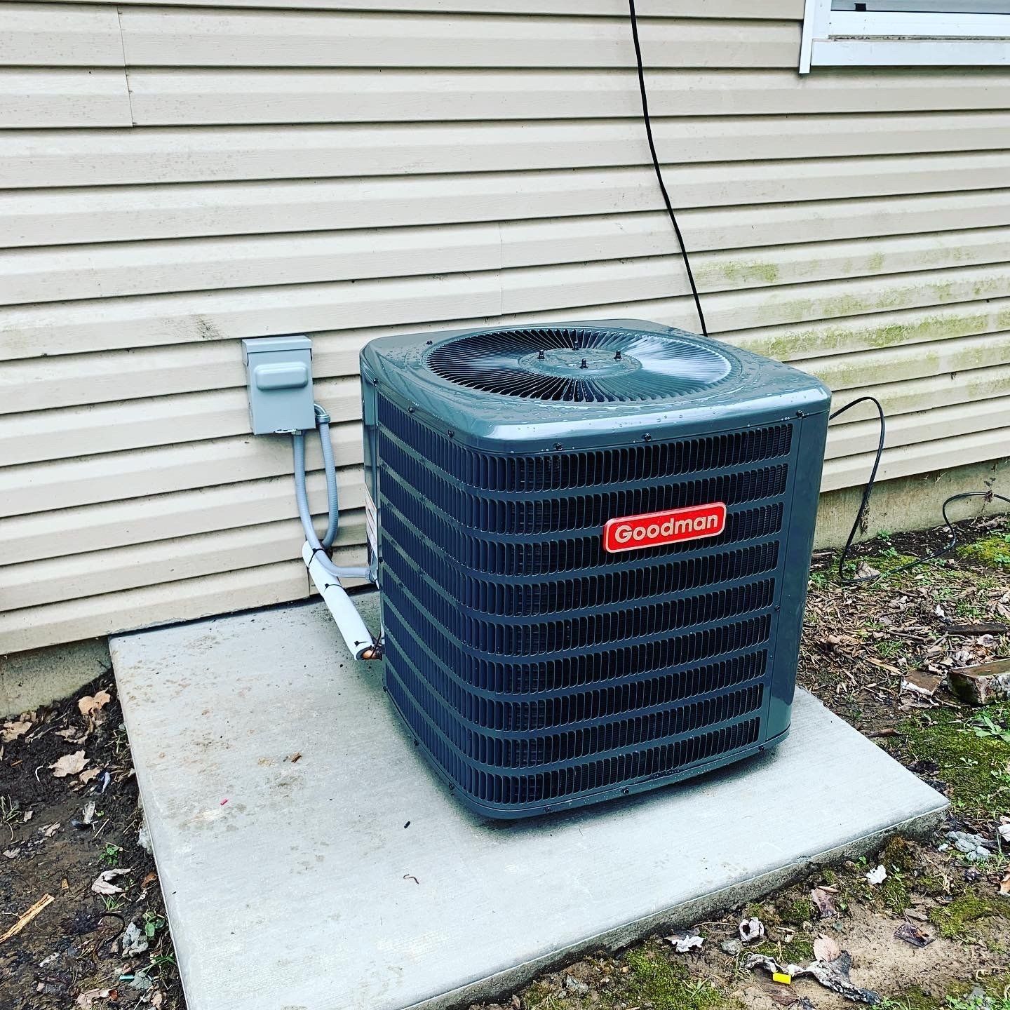 A green Goodman brand central air conditioning unit sits on a concrete slab next to beige house siding.