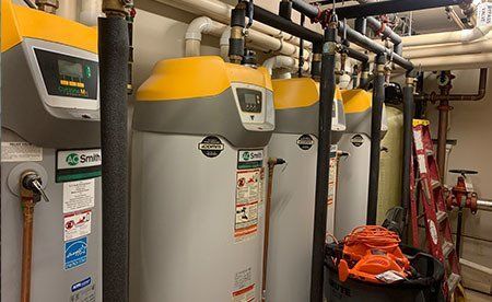 A row of four industrial A.O. Smith water heaters with yellow tops and digital displays in a mechanical room.