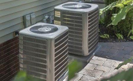 Two outdoor residential air conditioning units sit on a stone patio beside a house with light siding and brick.