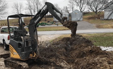 A John Deere mini excavator digs into a dirt pile on a rural lot next to a white pickup truck.