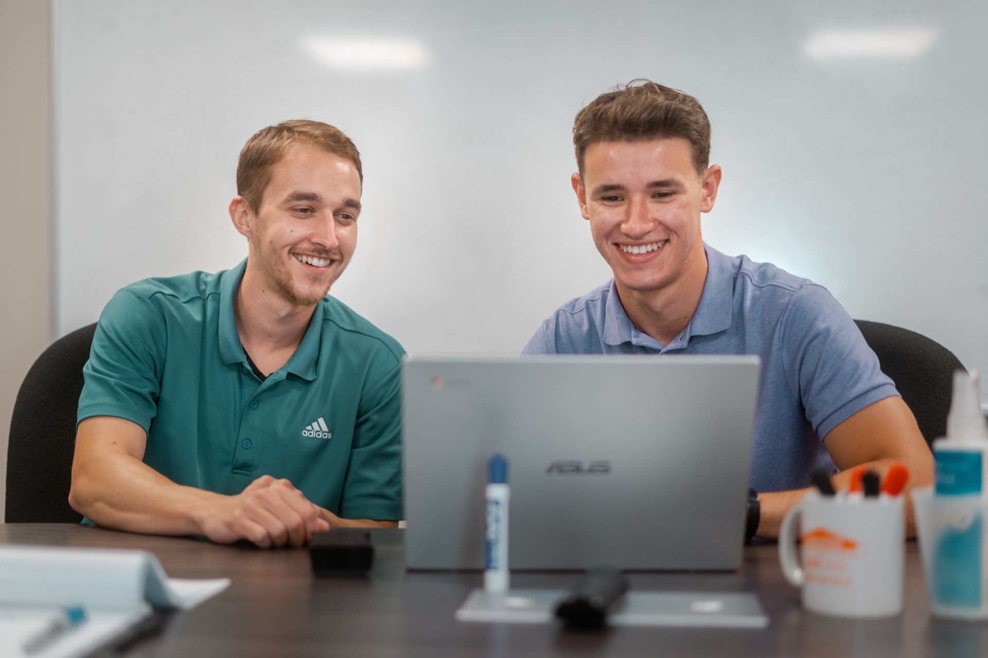 Two men are sitting at a table looking at a laptop computer.