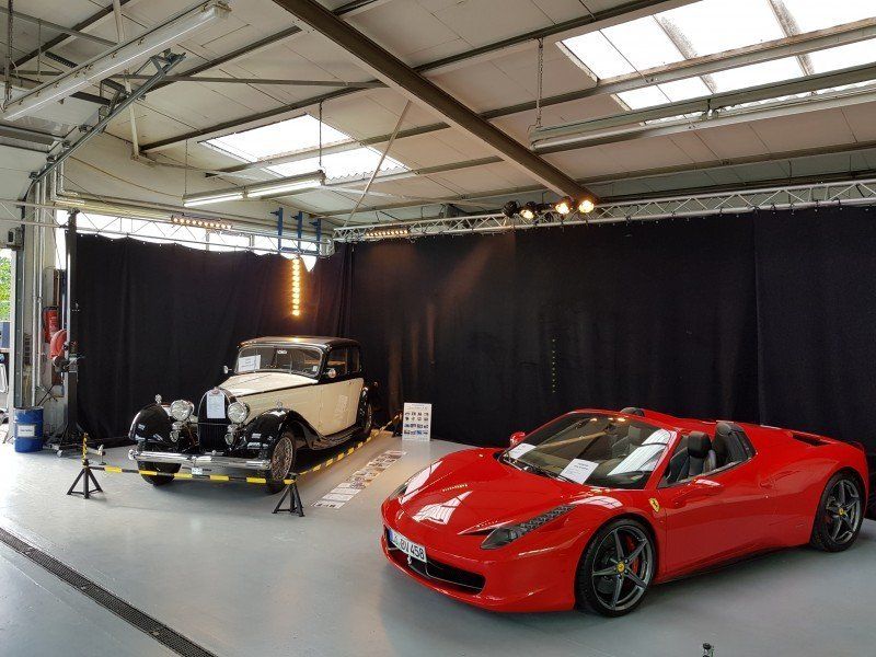 Ein roter Sportwagen und ein cremefarbener Oldtimer werden in einem hellen Ausstellungsraum präsentiert.