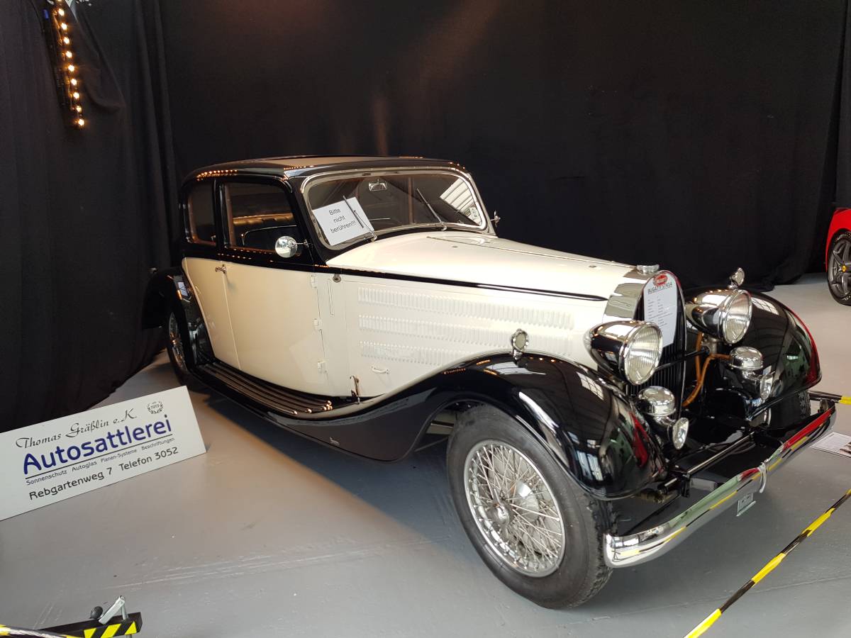 Ein klassischer, schwarz-cremefarbener Oldtimer ist in einem Innenraum ausgestellt, davor befindet sich ein Schild.