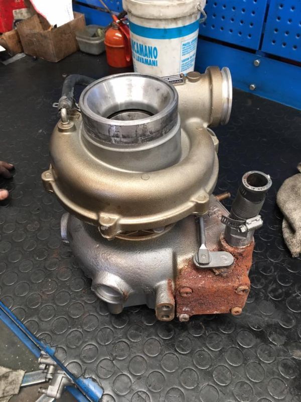 Un turbocompressore automobilistico in metallo è appoggiato su un banco da lavoro industriale scuro e decorato, accanto a un secchio bianco e ad alcuni componenti metallici.