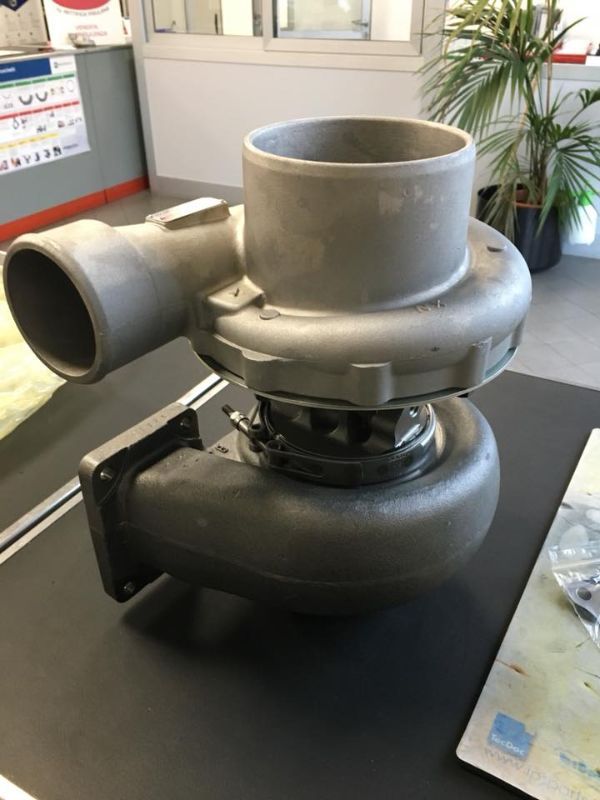 In un'officina, su una superficie nera, si trova un gruppo turbocompressore composto da un compressore in alluminio argentato e un alloggiamento in ferro.