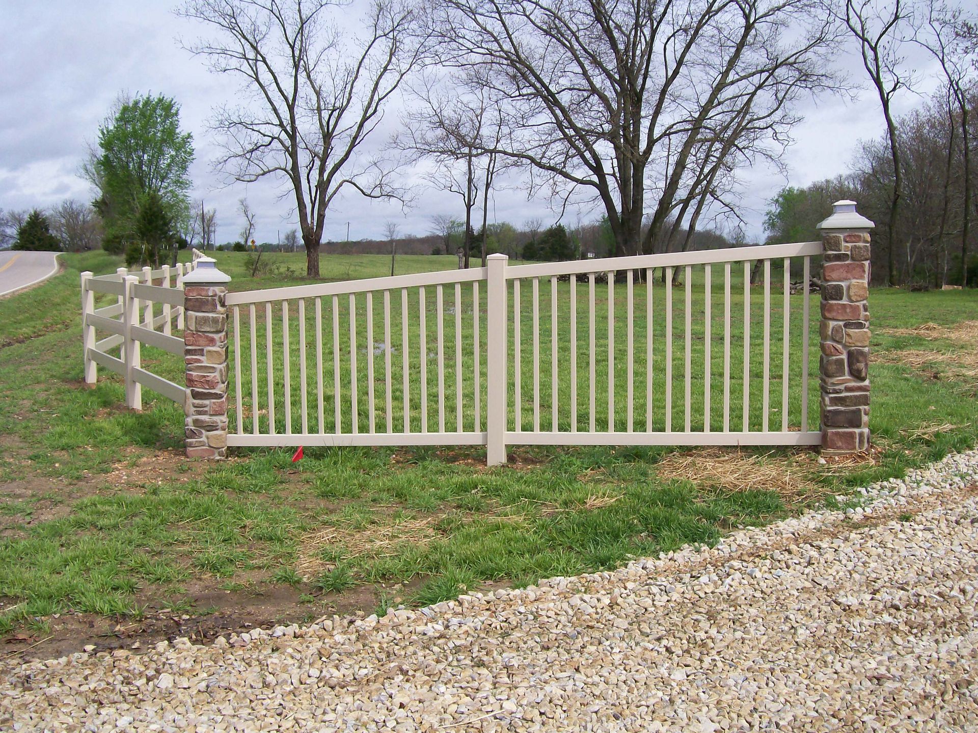 Beige and brown fence with stone pillars on grassy area, gravel driveway in front.