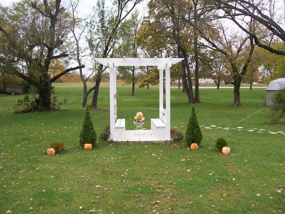 White arbor with benches and pumpkins in a grassy yard, surrounded by trees.