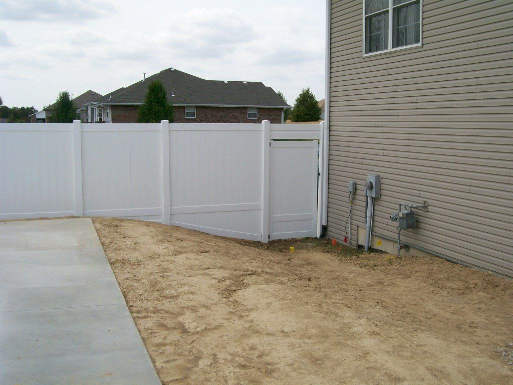 White vinyl fence with door, next to a beige house, bordering a sandy area, and concrete.