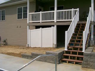 Exterior view of a raised deck with stairs. White railing, brown steps, beige house siding, and a white fence are visible.