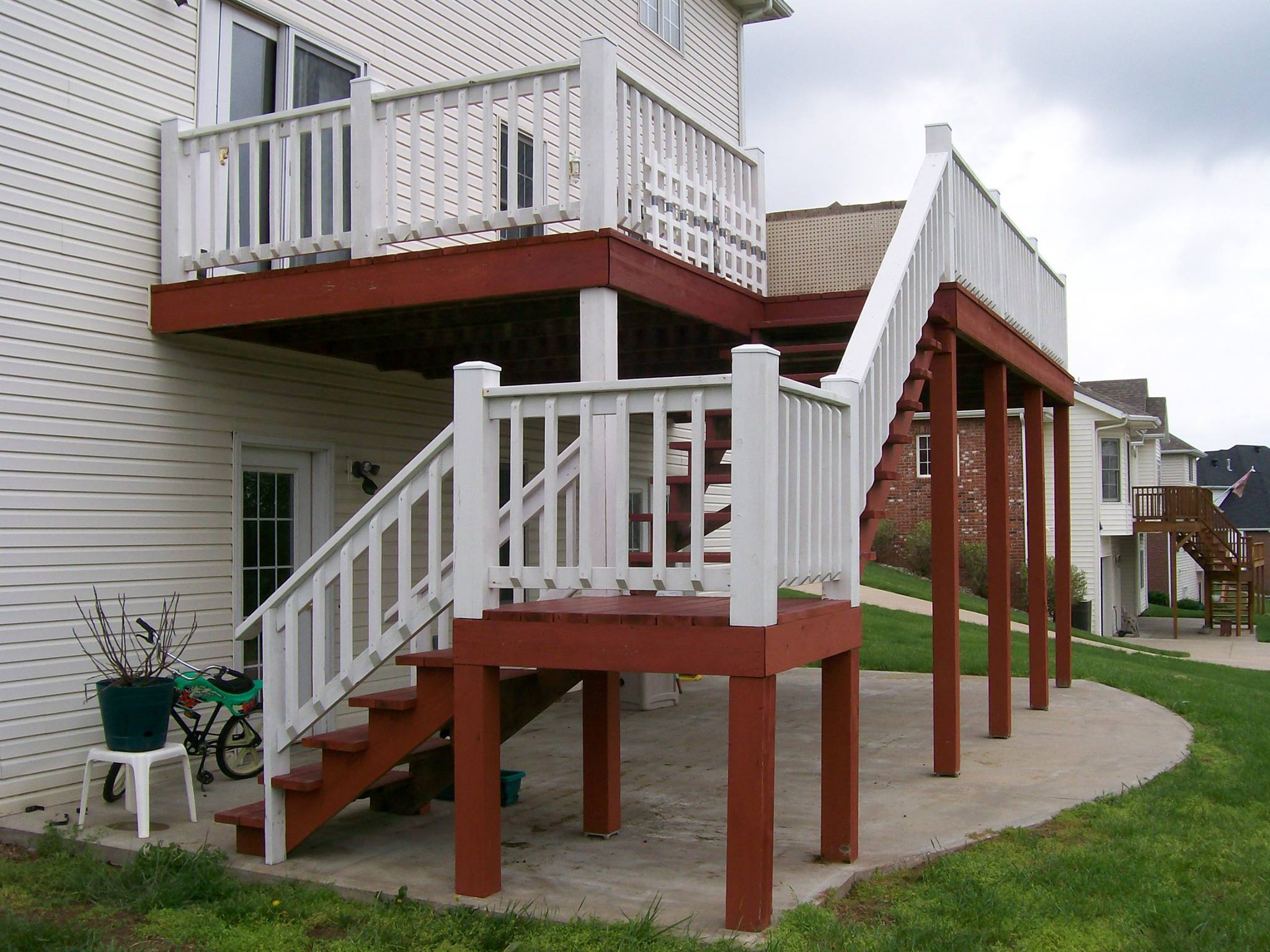 Two-story deck attached to a house with red and white railings, steps, and supports on a concrete patio.