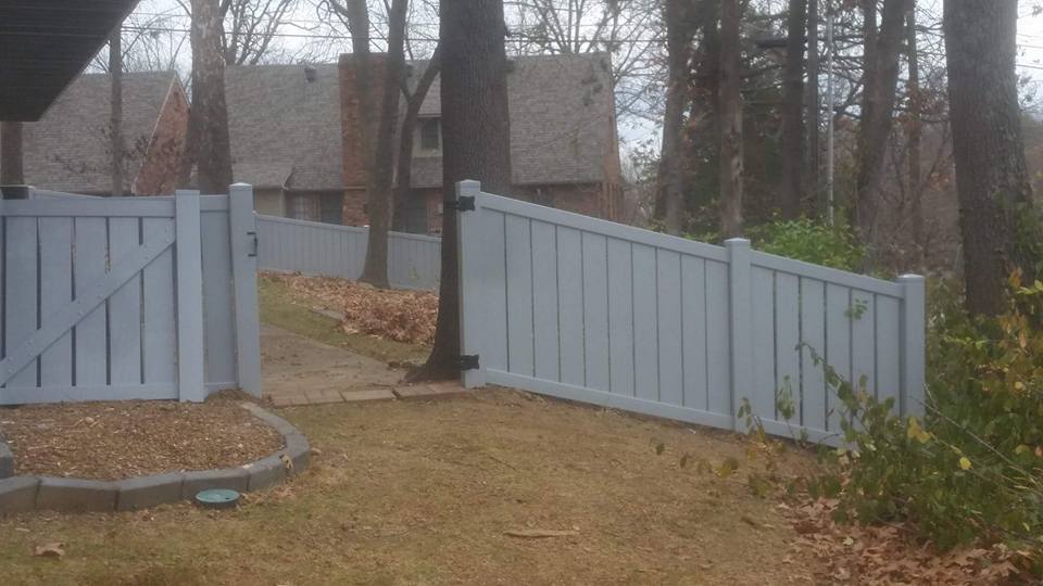 A light blue fence with a gate, trees and houses in the background. The grass is brown.