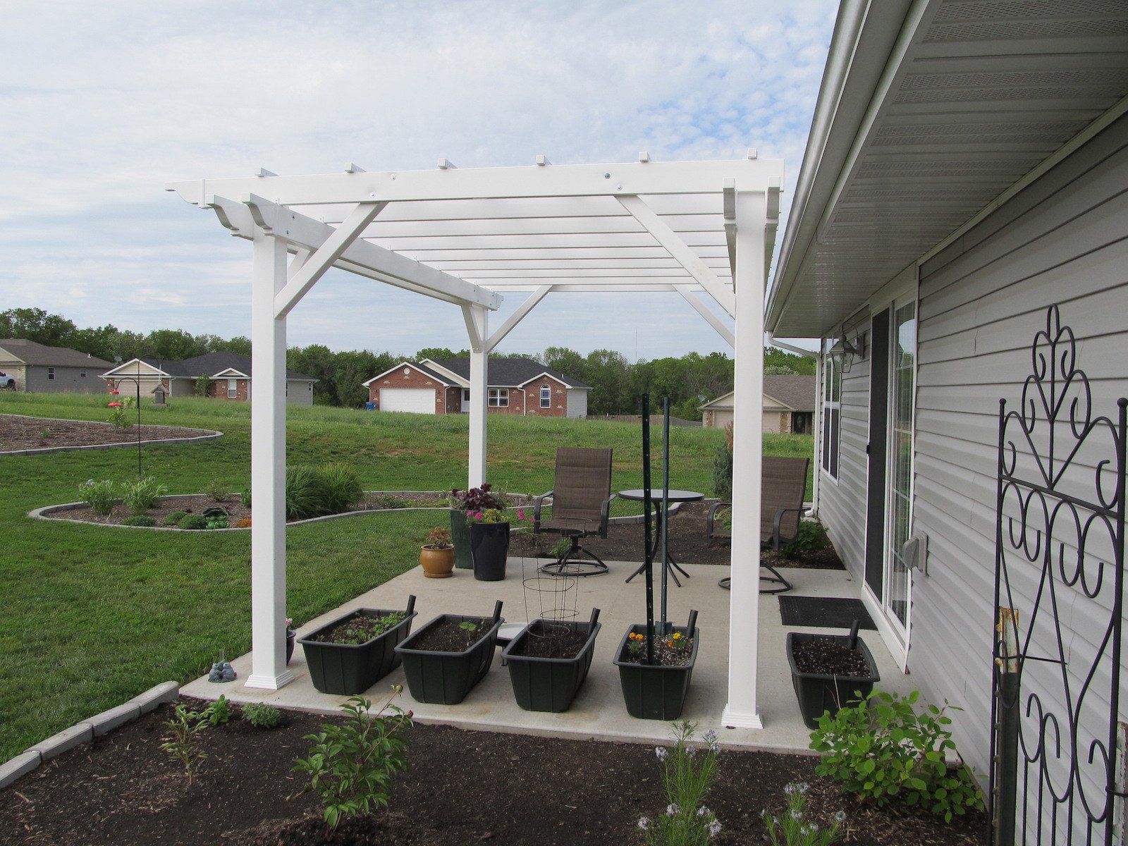 White pergola on a concrete patio with potted plants and yard, beside a house with sliding glass door.